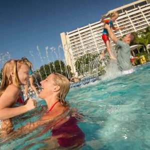 Photo of a woman throwing a young girl in the air amidst the beautiful blue green water of a pool. In the background a man and young boy splash. In the background, the mid-century modern a-frame design of Disney’s Contemporary Resort with its straight white lined architecture is framed by a clear blue sky.